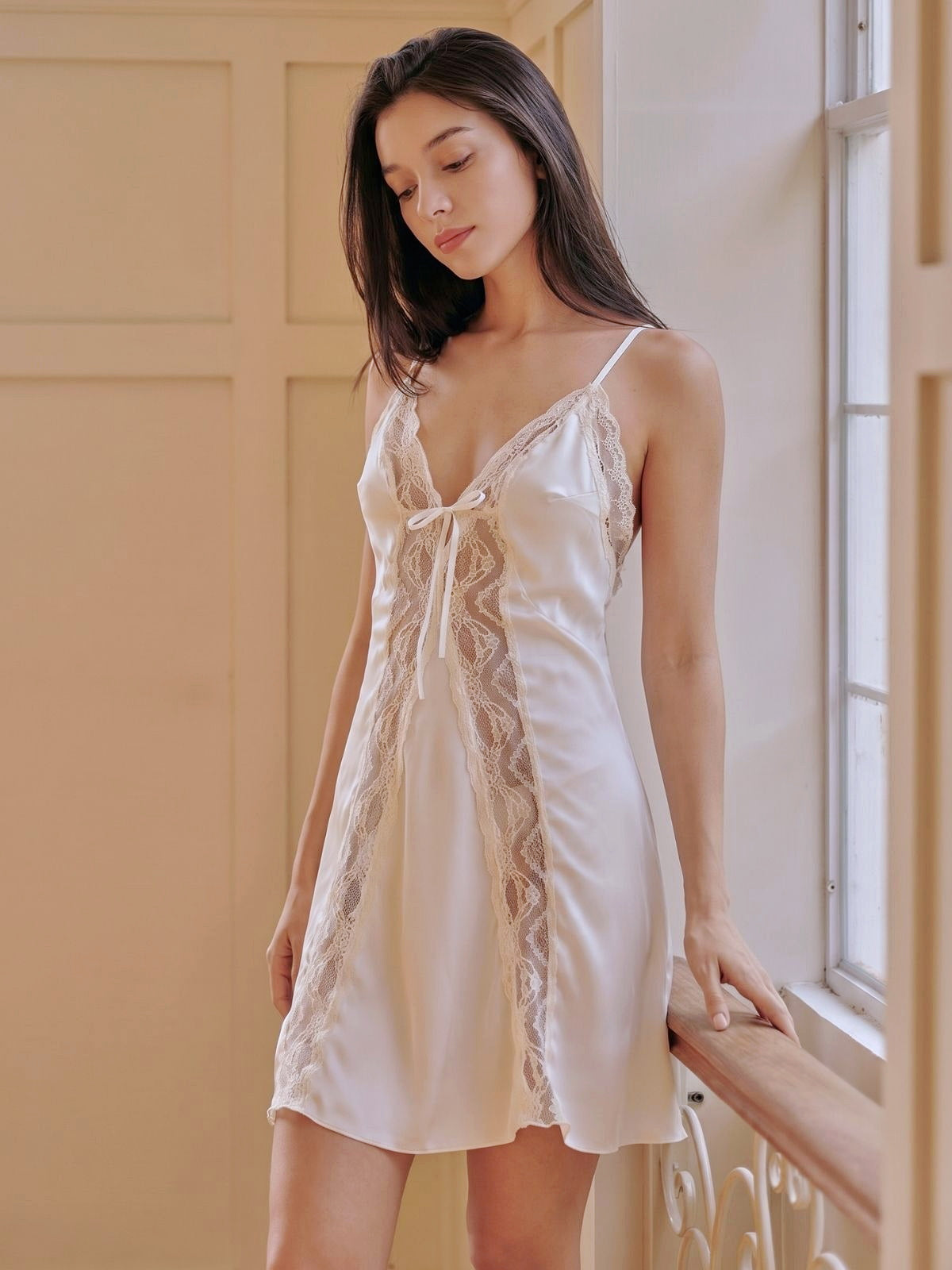Woman wearing a white lace nightgown standing by a window.