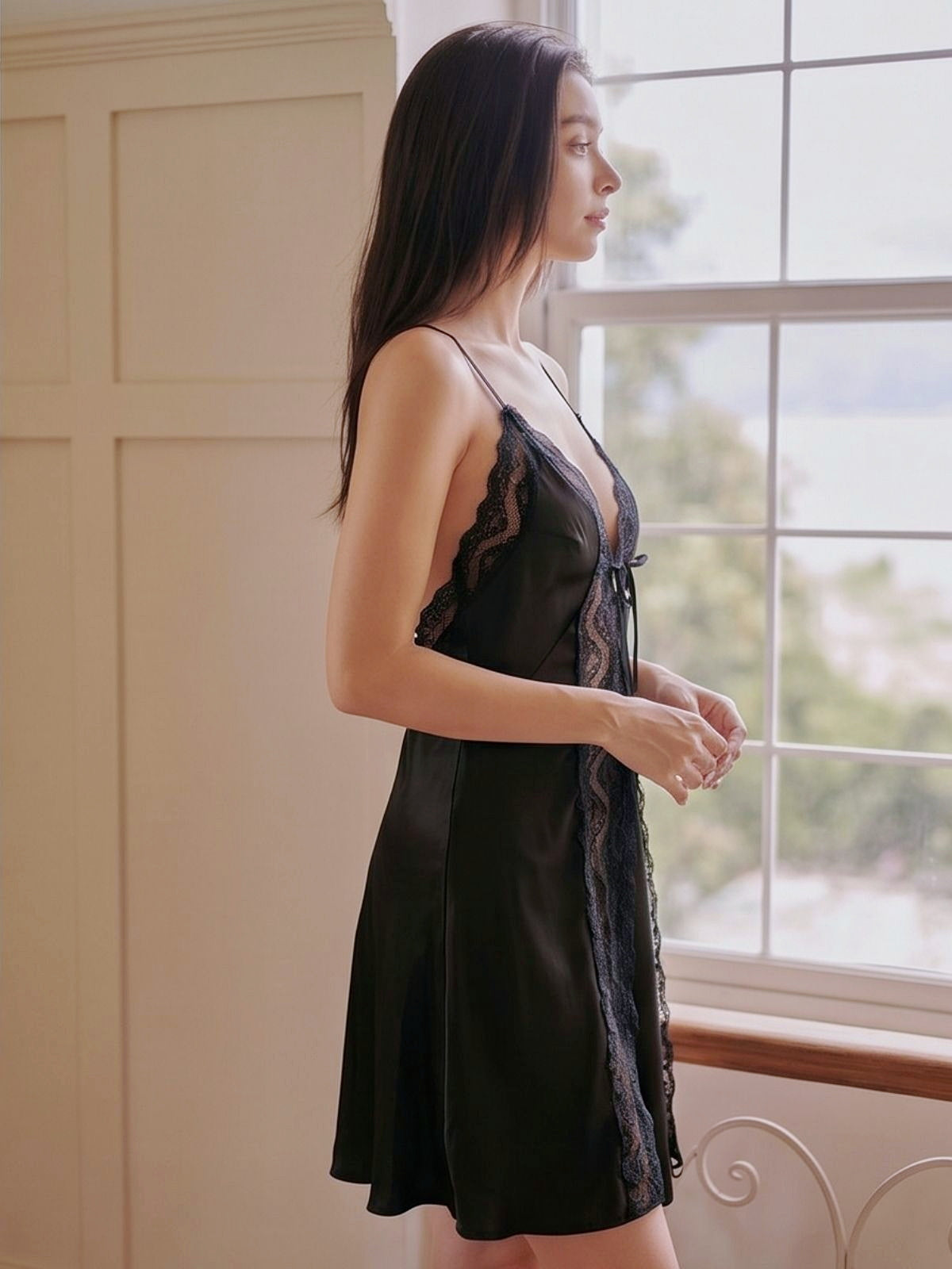 Woman wearing a black lace dress standing by a window.