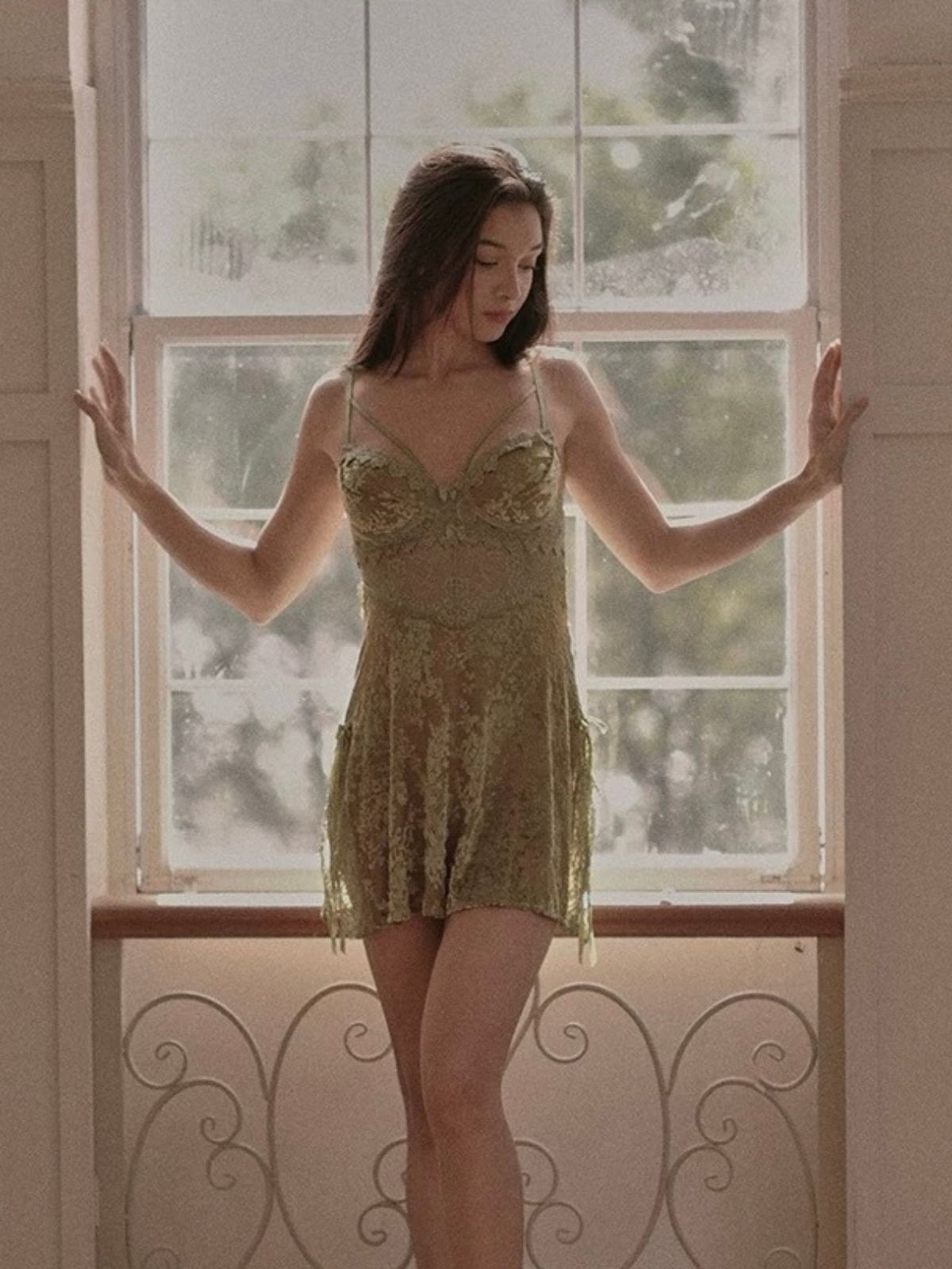 Woman in a green lace velvet material nightgown standing in front of a window with a blurred outdoor view.