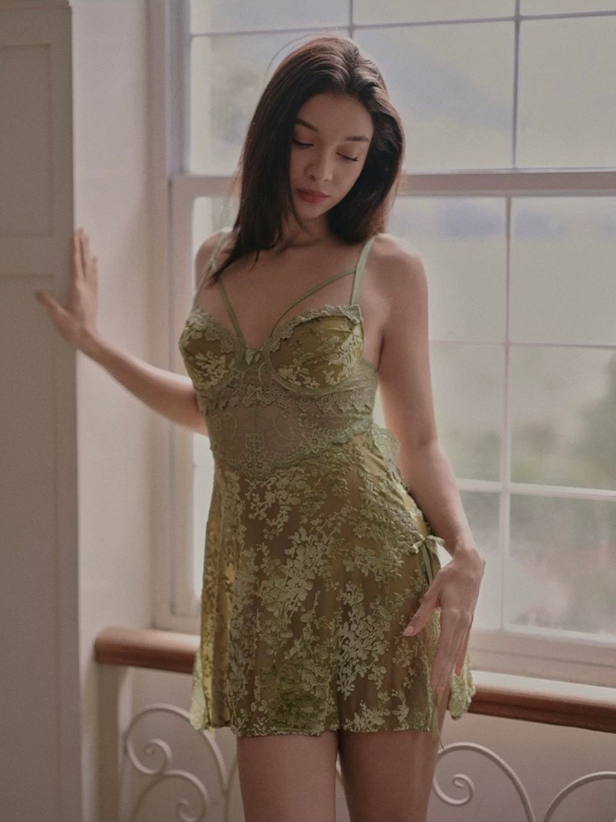 Woman wearing a green lace sleep dress standing in a room with large windows.