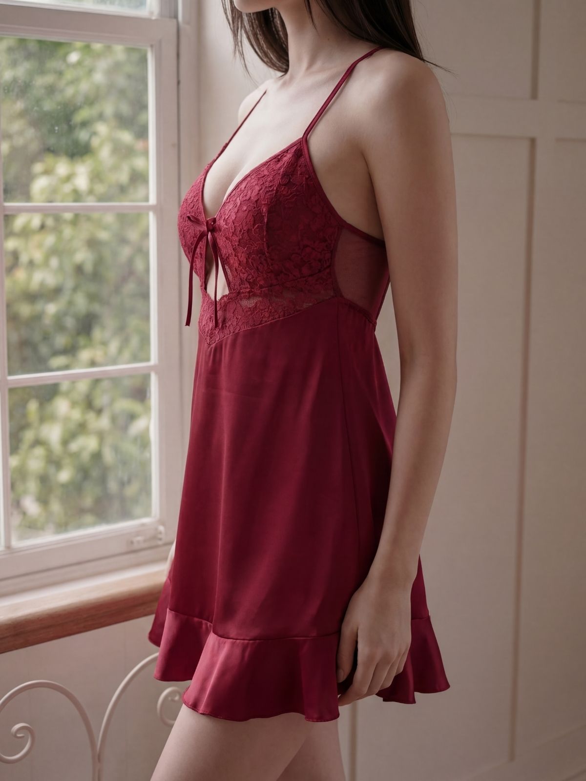 Woman wearing a red lace dress standing by a window with greenery outside.